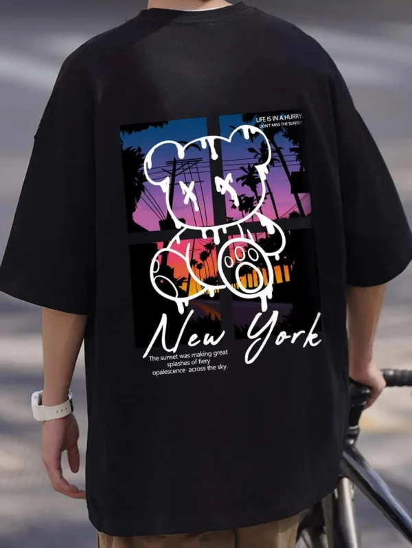 Men's loose fit cotton t-shirt displaying a colorful graphic print of a cartoon bear in front of a New York sunset scene.